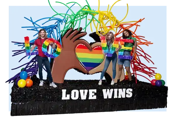 Pride parade float with large rainbow heart, colorful spray accents, and bold Love Wins lettering on a black fringe base.