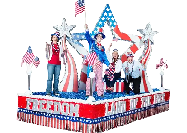 Patriotic parade float featuring people waving American flags, red white and blue balloons, and a large Freedom arch
