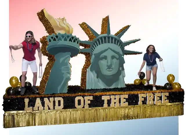 Patriotic parade float with oversized Statue of Liberty sculptures, gold fringe trim, and Land of the Free text across the front.