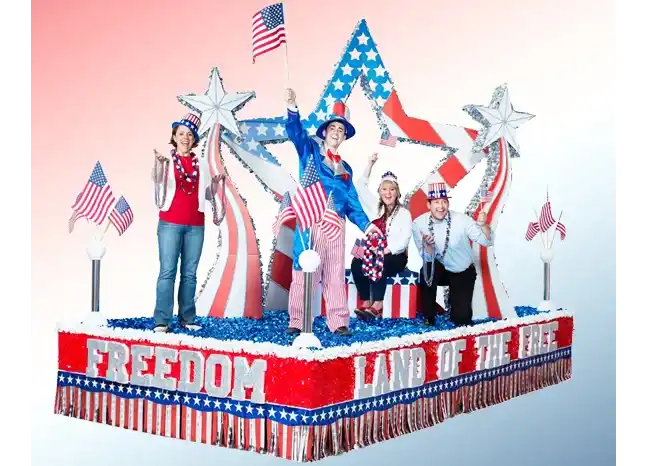 Patriotic parade float with people waving American flags, star decorations, and a large red white and blue Freedom display