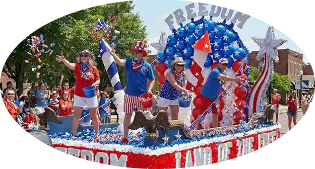 Patriotic parade float featuring people waving American flags, red white and blue balloons, and a large Freedom arch